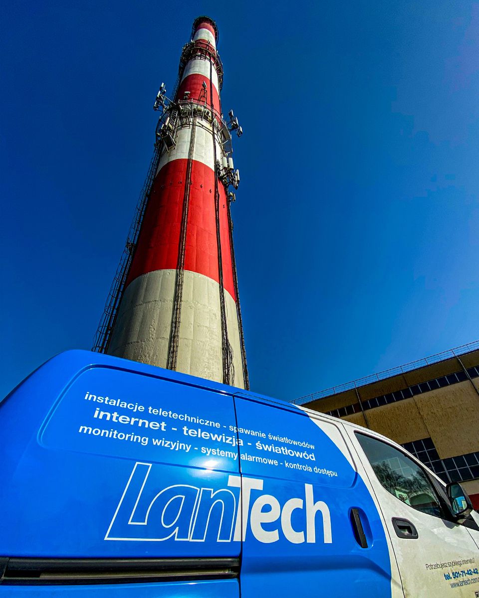 Niebiesko-białe auto LanTech z listą usług na boku: instalacje teletechniczne, internet, telewizja, światłowód, monitoring, alarmy, kontrola dostępu; w tle komin z antenami.