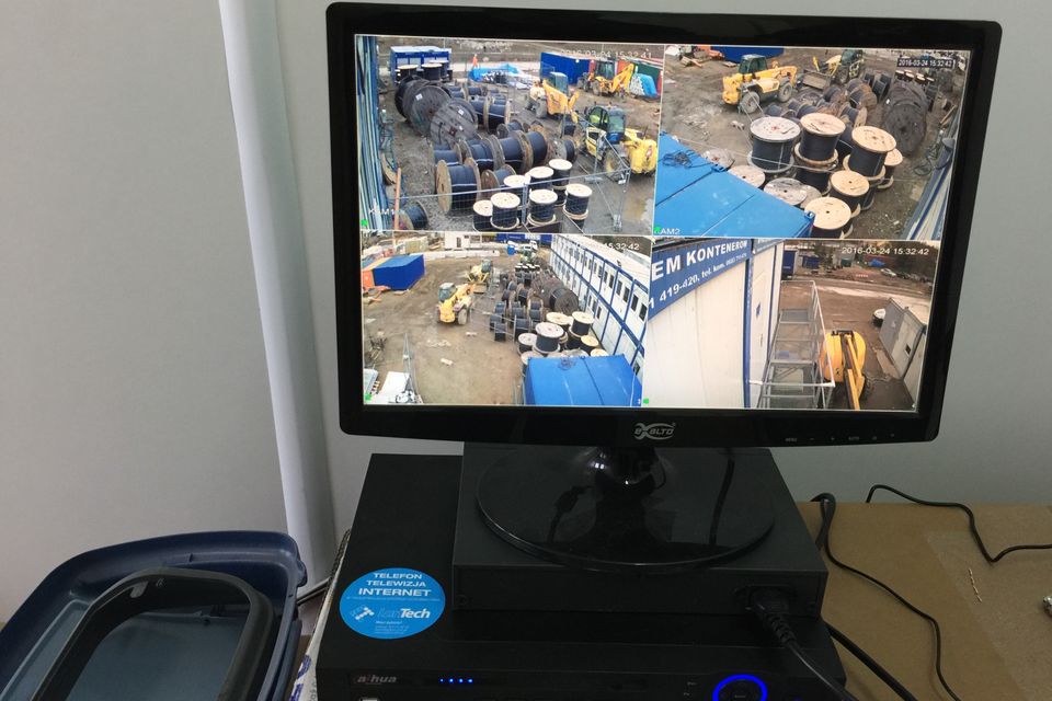 Monitor z podglądem czterech kamer CCTV placu budowy (szpule kabli, ładowarki teleskopowe i kontenery) stojący na rejestratorze z naklejką LanTech.