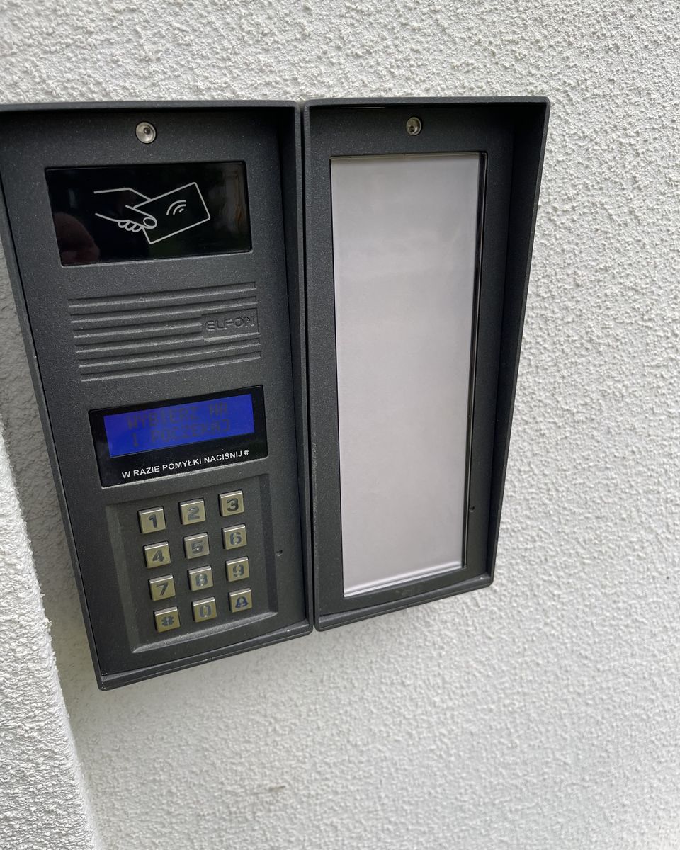 Panel domofonowy na elewacji z klawiaturą, wyświetlaczem i polem czytnika breloków, obok pusta tablica.