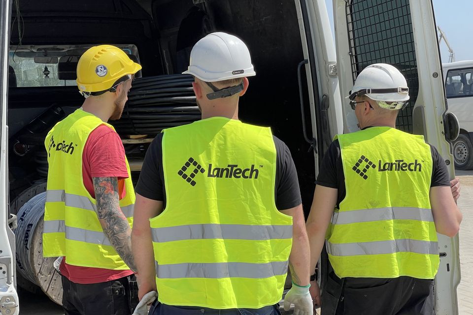Trzech monterów w kaskach i kamizelkach z logo LanTech, odwróconych tyłem, przy otwartym aucie z bębnami kabla.