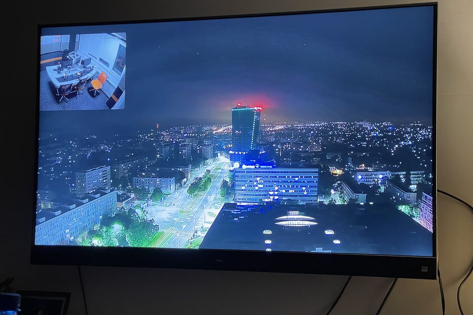 Telewizor na ścianie w pomieszczeniu bez okien: nocna panorama miasta z kamery na żywo, w lewym górnym rogu mniejszy obraz ze studia.