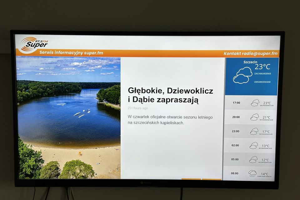 Ekran przy wejściu do biurowca z serwisem informacyjnym radia Super FM: zdjęcie z artykułu, nagłówek i prognoza pogody dla Szczecina na kolejne godziny.