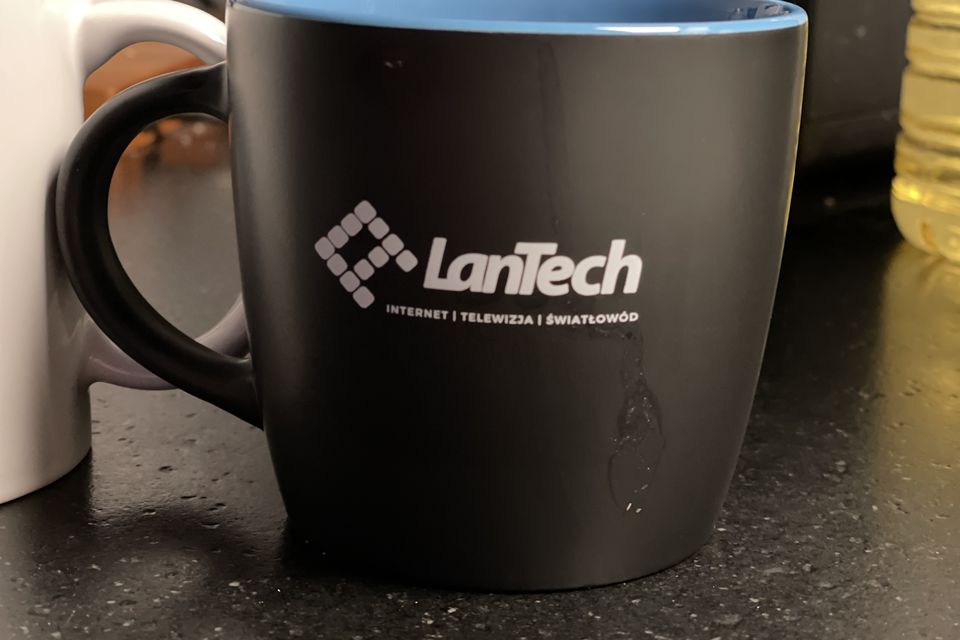 Czarny kubek z niebieskim wnętrzem i logo LanTech z dopiskiem internet, telewizja, światłowód, na blacie w biurze.