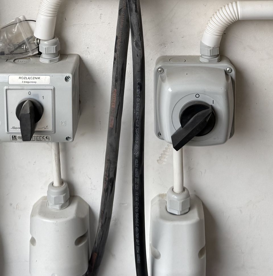 Dwa rozłączniki trójbiegunowe w obudowach natynkowych, peszle, dławiki i przemysłowe gniazda zasilające na ścianie garażu.