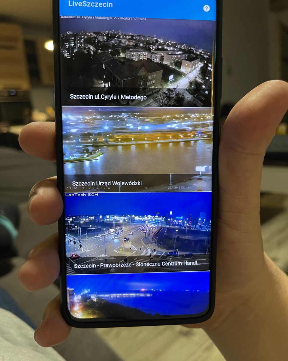 Telefon w dłoni z aplikacją LiveSzczecin pokazującą kafelki kamer na żywo z widokami nocnego miasta.