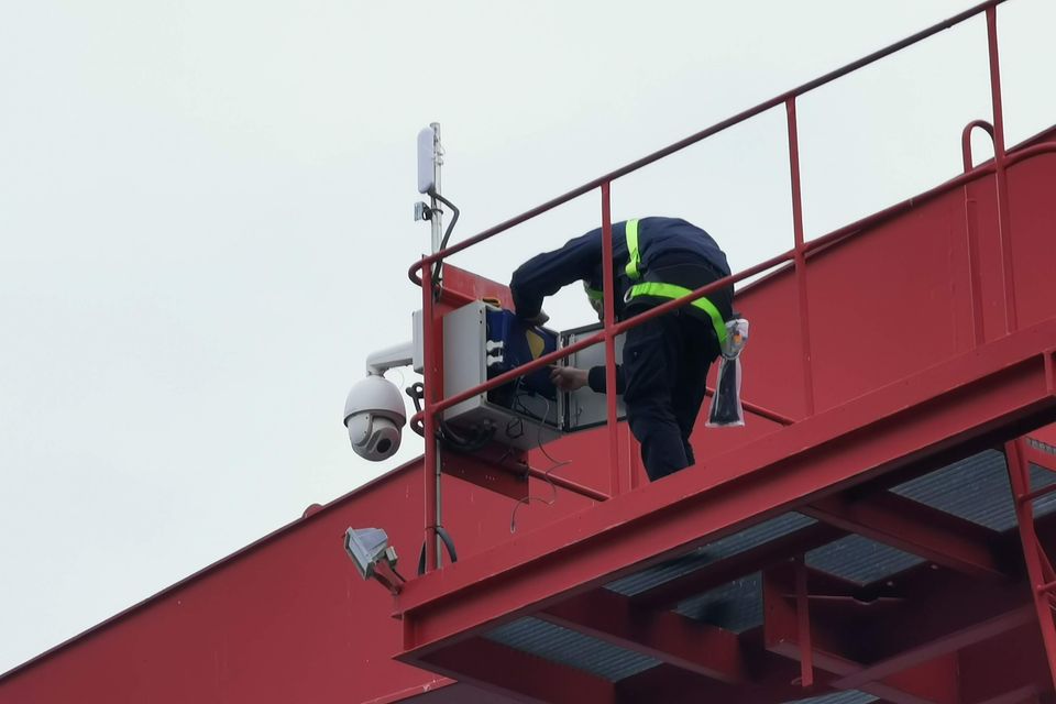 Technik w uprzęży przy skrzynce na czerwonej konstrukcji; obok obrotowa kamera CCTV i antena łącza radiowego.