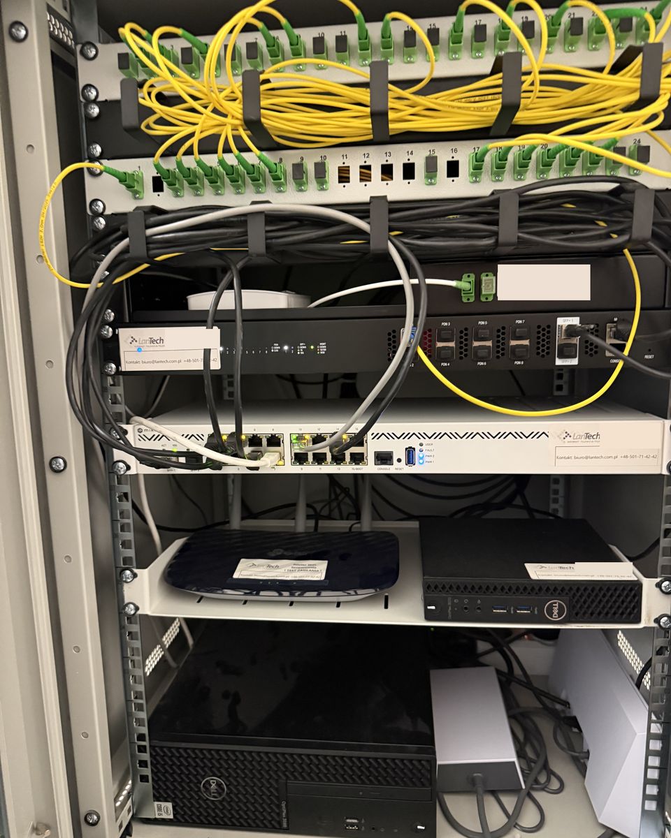 Szafa sieciowa obiektu: przełącznica światłowodowa z żółtymi patchcordami, zakończenie łącza, router z etykietą LanTech i sprzęt na dolnych półkach.