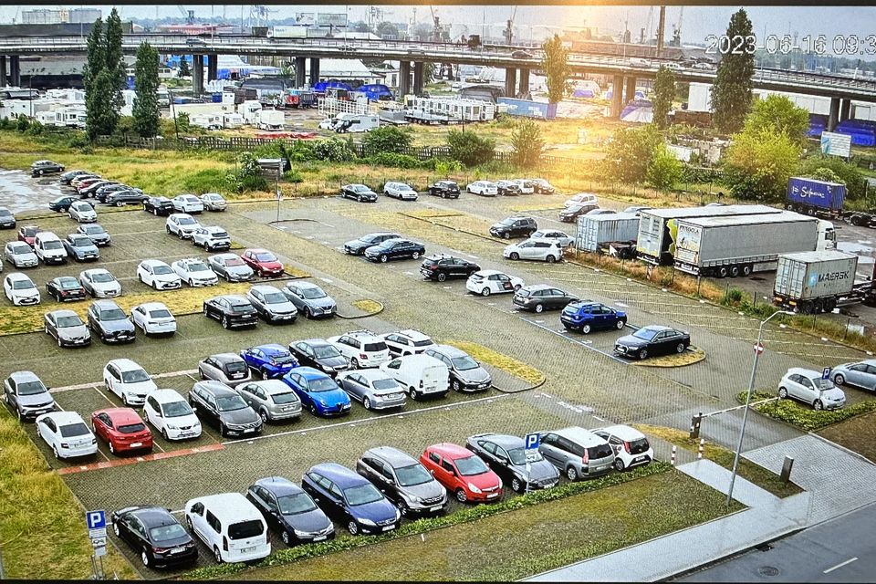 Obraz z kamery CCTV zawieszonej wysoko nad dużym parkingiem: rzędy samochodów, wolne miejsca, wjazd i ciężarówki w tle.