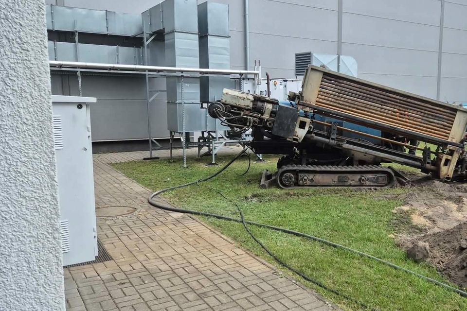 Wiertnica do przewiertu sterowanego ustawiona na trawniku przy ścianie hali, z przewodami prowadzącymi do wykopu.