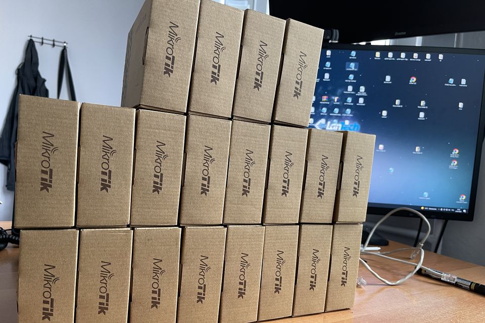 Stos kilkudziesięciu routerów w kartonach na biurku obok monitora.