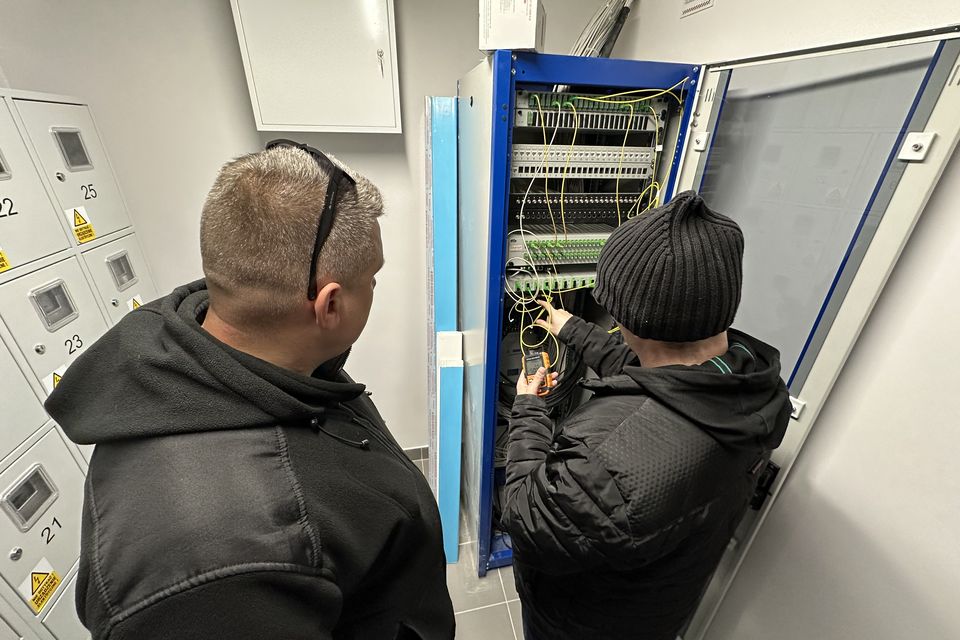 Dwóch techników LanTech, widocznych od tyłu, przy otwartej szafie światłowodowej na klatce schodowej; jeden mierzy tor ręcznym miernikiem.
