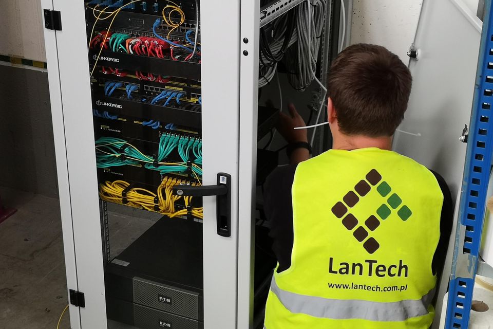 Technik w kamizelce z logo LanTech, odwrócony tyłem, pracuje przy nowej szafie teletechnicznej z opisanym okablowaniem.