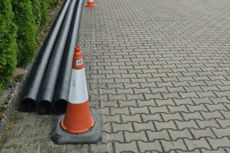 Wiązka czarnych rur osłonowych ułożona wzdłuż krawędzi drogi z kostki, oddzielona pachołkami od ruchu.