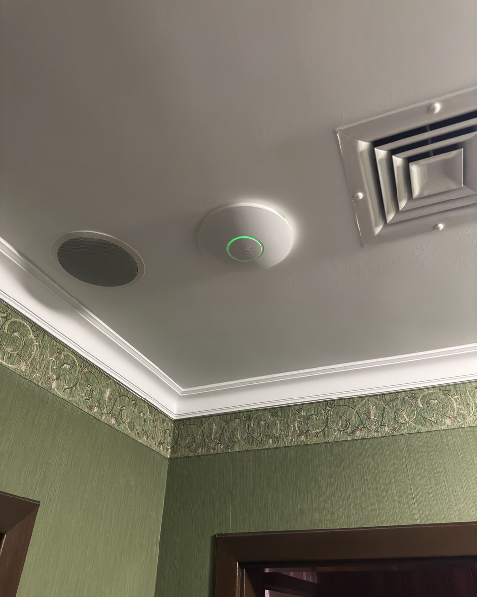 Punkt dostępowy Wi-Fi z zieloną diodą na suficie pokoju hotelowego, obok kratka wentylacyjna.